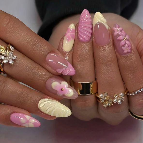 A close-up of manicured hands wearing long almond-shaped press-on nails in pastel pink, cream, and lavender, with floral and shell designs, along with gold rings.