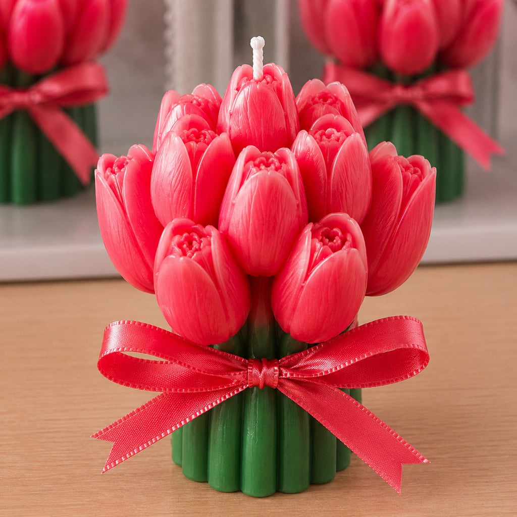 A tulip-shaped candle with bright pink-red petals, green stems, and a red ribbon bow, shown on a wooden surface with blurred matching candles in the background.