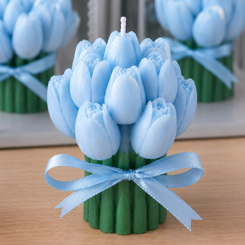 A tulip-shaped candle with pale blue petals and green stems, tied with a matching light blue ribbon, shown on a wooden surface with blurred mirrors in the background.