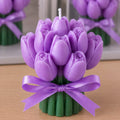 A purple tulip-shaped candle with a white wick, tied with a purple satin ribbon and green fluted base, shown on a wooden surface with blurred matching candles in the background.
