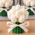 A tulip-shaped candle made of white wax with a green stem base and a cream ribbon bow, shown on a wooden surface with a softly blurred mirror reflection behind it.